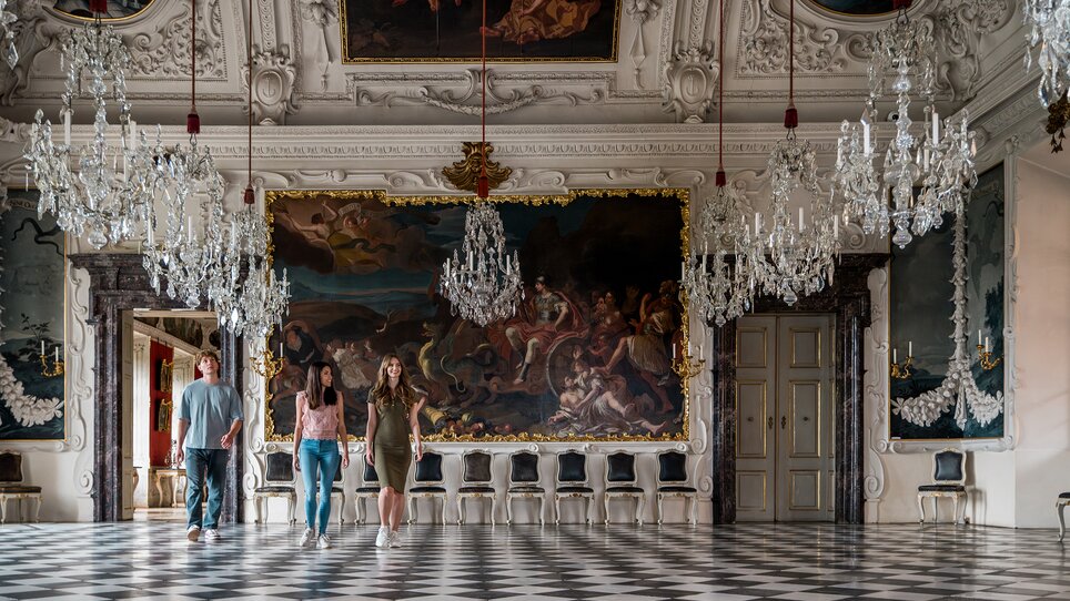 Drei Personen gehen durch einen opulenten Raum mit Kristallleuchtern und Wandmalerei im Schloss Eggenberg. | © Graz Tourismus - Mias Photoart