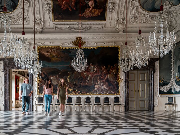 Tre persone attraversano una sala opulenta con lampadari di cristallo e pitture murali nel Palazzo Eggenberg. | © Graz Tourismus - Mias Photoart