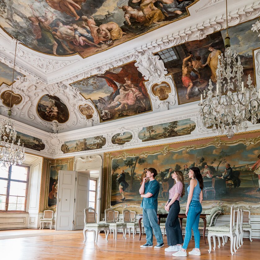 Three people walk through an opulent room with crystal chandeliers and wall paintings in Eggenberg Palace. | © Graz Tourismus - Mias Photoart