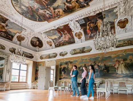 Drei Personen gehen durch einen opulenten Raum mit Kristallleuchtern und Wandmalerei im Schloss Eggenberg. | © Graz Tourismus - Mias Photoart