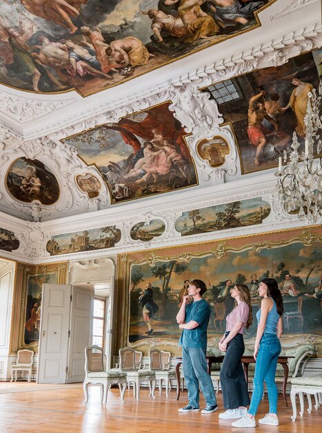 Drei Personen gehen durch einen opulenten Raum mit Kristallleuchtern und Wandmalerei im Schloss Eggenberg. | © Graz Tourismus - Mias Photoart