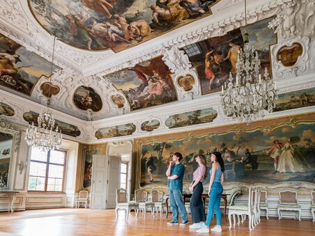 Drei Personen gehen durch einen opulenten Raum mit Kristallleuchtern und Wandmalerei im Schloss Eggenberg. | © Graz Tourismus - Mias Photoart