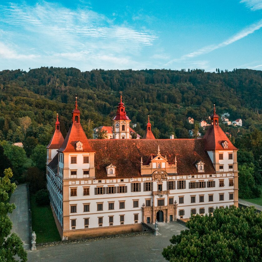Castello di Eggenberg a Graz | © Graz Tourismus - Mias Photoart Una vista del Castello di Eggenberg, circondato da alberi e montagne. | © Graz Tourismus - Mias Photoart