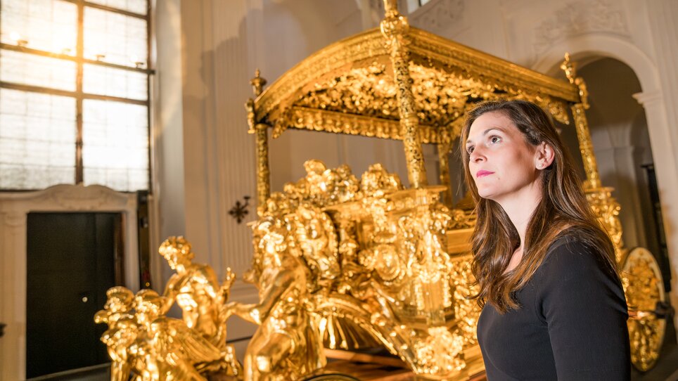 Eine Frau steht vor einem kunstvoll verzierten goldenen Prunkwagen. | © Graz Tourismus - Mias Photoart