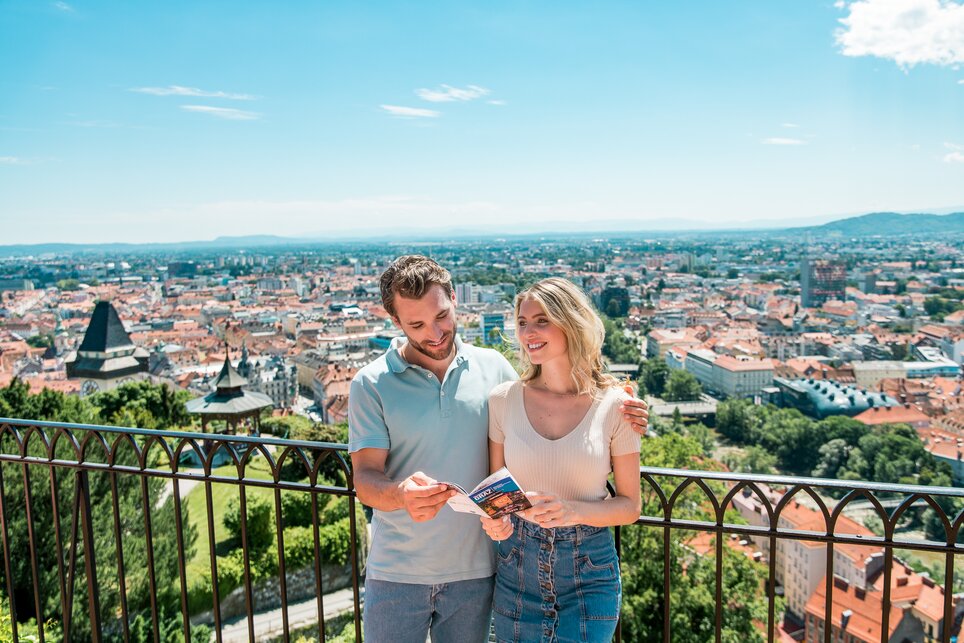 Paar genießt den Ausblick über Graz vom Schlossberg. Die beiden betrachten einen Prospekt von Graz. | © Graz Tourismus - Mias Photoart