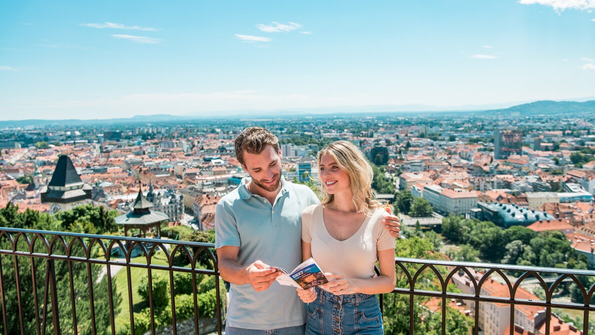 Couple enjoying the view over Graz from the Schlossberg. The two are looking at a brochure of Graz. | © Graz Tourismus - Mias Photoart