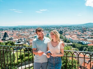 Paar genießt den Ausblick über Graz vom Schlossberg. Die beiden betrachten einen Prospekt von Graz. | © Graz Tourismus - Mias Photoart