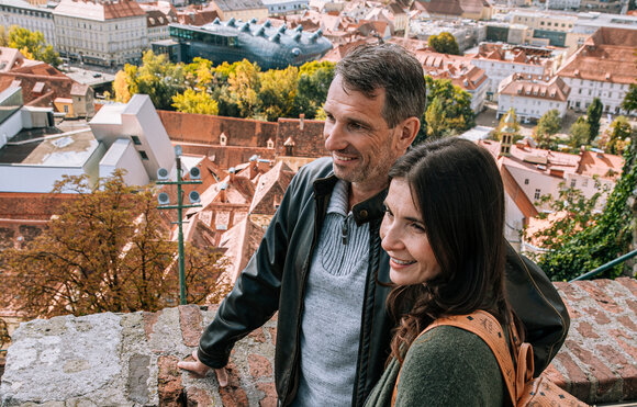 Paar genießt die Aussicht auf Graz. Im Hintergrund ist das Kunsthaus und die wunderschöne rote Dachlandschaft zu sehen. | © Graz Tourismus