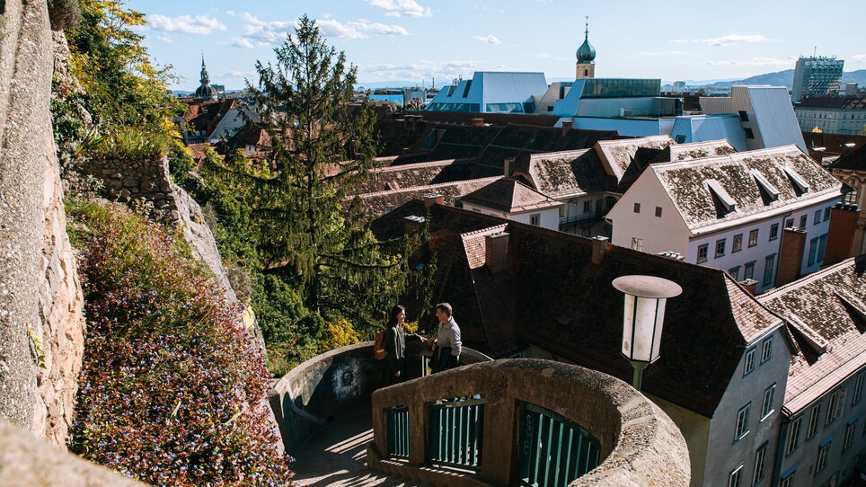 View of Graz from the Schlossbergsteig with red rooftops in the background. A couple is walking up the Schlossbergsteig. | © Graz Tourismus