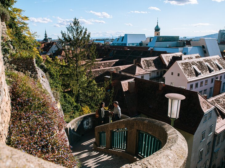 Veduta su Graz dallo Schlossbergsteig con i tetti rossi sullo sfondo. Una coppia sta salendo lo Schlossbergsteig. | © Graz Tourismus