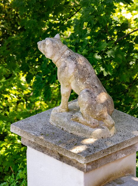 Una scultura di pietra di un cane su un piedistallo, circondata da fogliame verde. | © Graz Tourismus - Harry Schiffer
