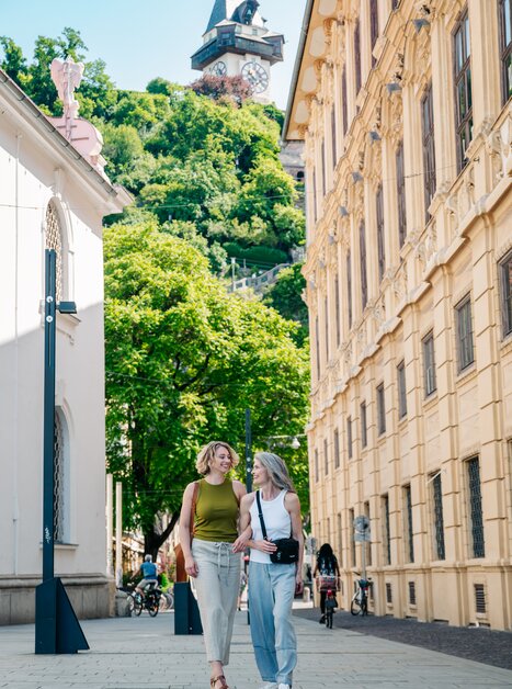 Due donne che passeggiano a Graz con la Torre dell'Orologio di Graz sullo sfondo. | © Graz Tourismus - Mias Photoart