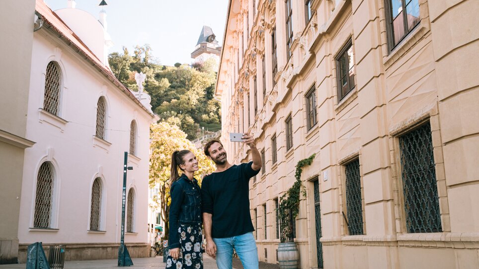 Ein Paar macht ein Selfie in der Stadt Graz, mit dem Grazer Uhrturm im Hintergrund. | © Graz Tourismus