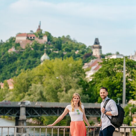Coppia vicino alla Mur con Torre dell'Orologio di Graz sullo sfondo. | © Graz Tourismus - Mias Photoart