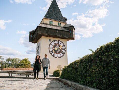 Paar spaziert vor dem Grazer Uhrturm bei blauem Himmel. | © Graz Tourismus