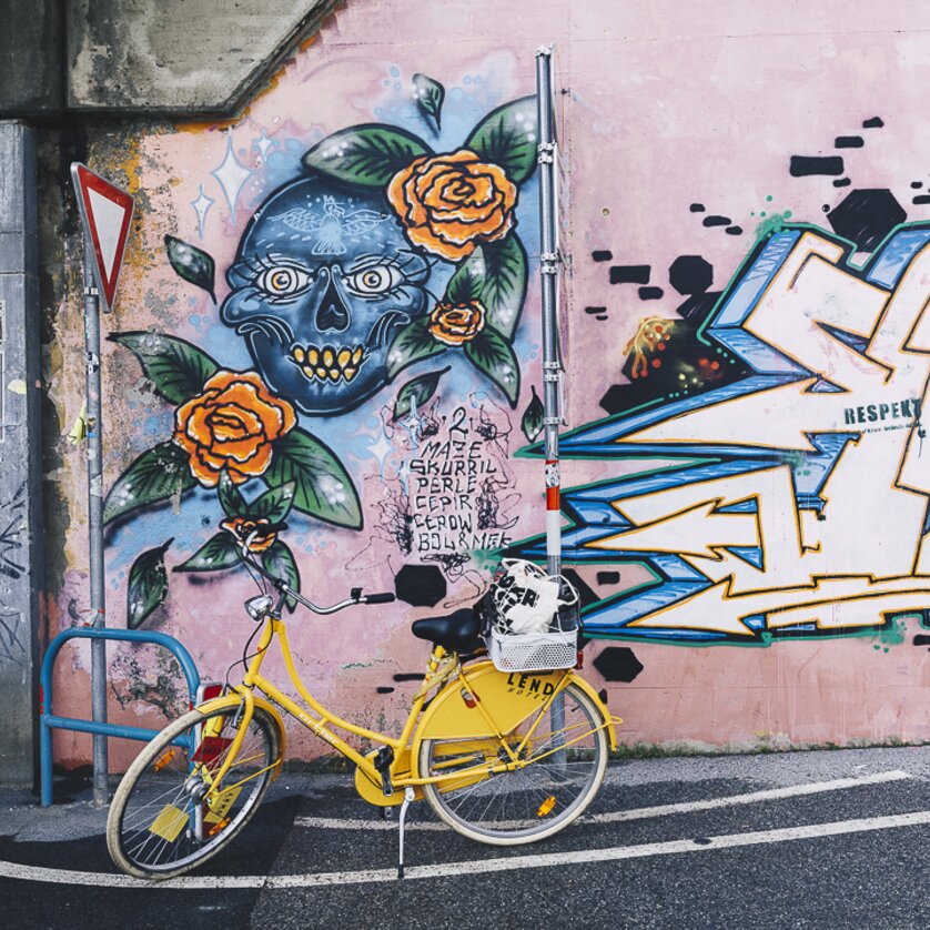 Graffiti-Kunst in Graz | © Adeline Gressin Gelbes Fahrrad vor einer Wand mit farbenfrohem Graffiti von einem Totenkopf und Rosen. | © Adeline Gressin