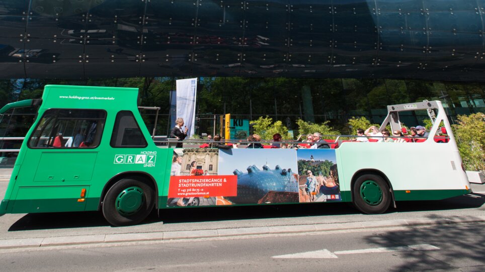 Ein Cabriobus fährt am Kunsthaus Graz vorbei, blauer Himmel und grüne Bäume im Hintergrund. | © Graz Tourismus - Harry Schiffer