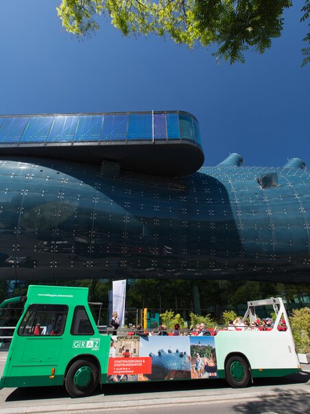 A cabrio bus passes by Kunsthaus Graz, blue sky and green trees in the background. | © Graz Tourismus - Harry Schiffer