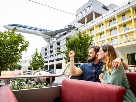 Coppia che si gode il panorama durante un tour culinario in un autobus decappottabile - sullo sfondo un aereo del ristorante NOVAPARK. | © Graz Tourismus - Werner Krug