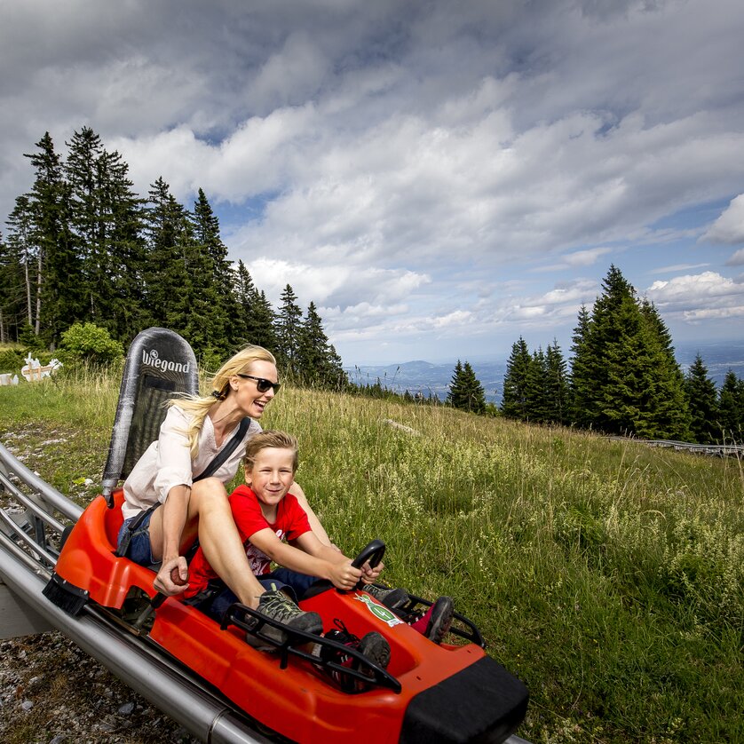 Summer Toboggan Run Hexenexpress Schöckl | © Region Graz - Tom Lamm A mother and her son ride a summer toboggan in the mountains. | © Region Graz - Tom Lamm