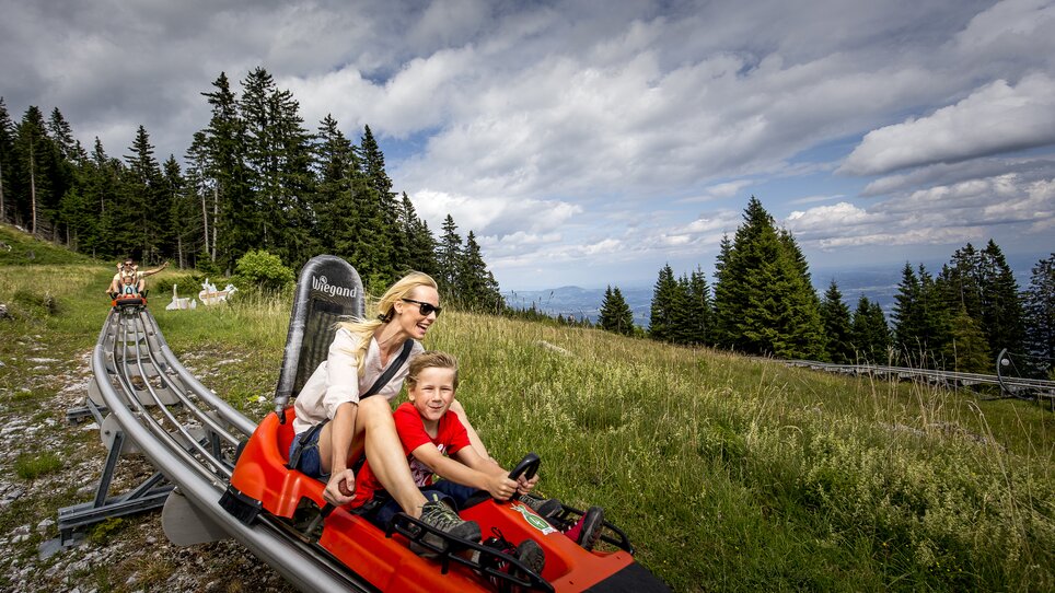 Eine Mutter und ihr Sohn fahren auf einer Sommerrodelbahn in den Bergen. | © Region Graz - Tom Lamm