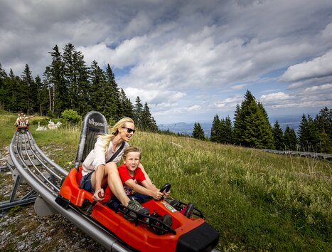 Eine Mutter und ihr Sohn fahren auf einer Sommerrodelbahn in den Bergen. | © Region Graz - Tom Lamm