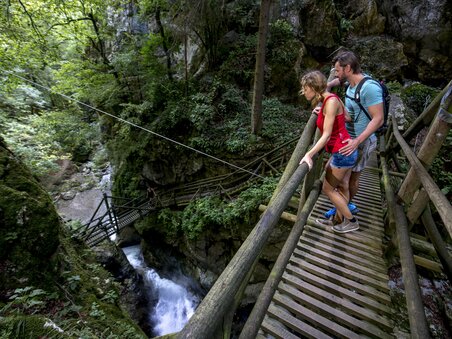 A couple stands on a wooden bridge overlooking the river in Kesselfallklamm. | © Region Graz - Tom Lamm