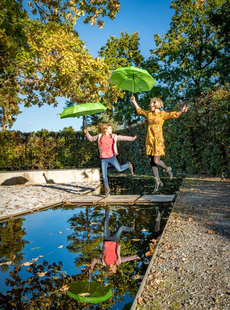 Ein Mädchen und eine Frau springen über eine Wasserfläche mit grünen Regenschirmen. | © Region Graz - Mias Photoart