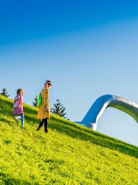 Eine Mutter und ihre Tochter spazieren durch den Österreichischen Skulpturenpark bei Graz, im Hintergrund ist der riesige Griff eines Koffers zu sehen. | © Region Graz - Mias Photoart