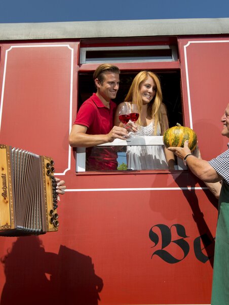 Flascherlzug Stainz | © Steiermark Tourismus - Harry Schiffer A couple on the Flascherlzug in Stainz receives wine and a pumpkin through the train window from a local farmer, while a woman in traditional dress plays a Styrian accordion on the platform. | © Steiermark Tourismus - Harry Schiffer