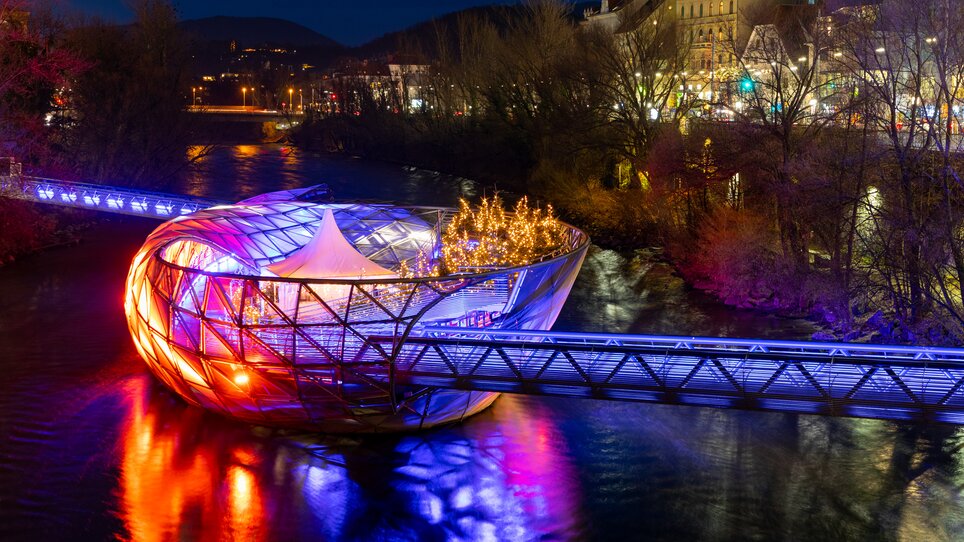 Mur Island in Graz shines with colorful lights above the Mur River. | © Graz Tourismus - Harry Schiffer I Lichtkonzept: Ochoresotto  