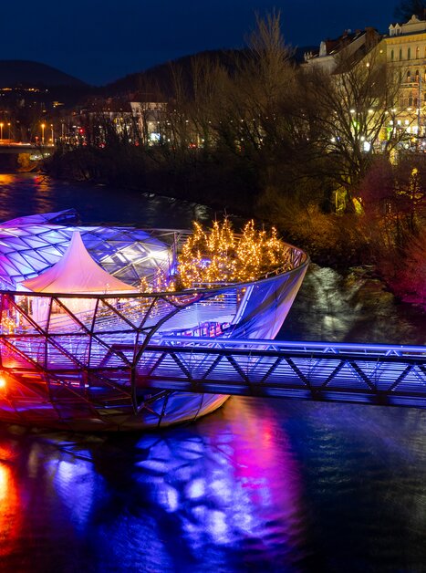 Mur Island in Graz shines with colorful lights above the Mur River. | © Graz Tourismus - Harry Schiffer I Lichtkonzept: Ochoresotto  