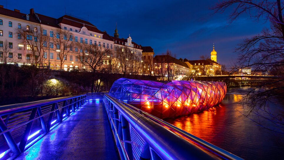 Illuminated Island in the Mur in Graz at night with a view of the city. | © Graz Tourismus - Harry Schiffer I Lichtkonzept: Ochoresotto  