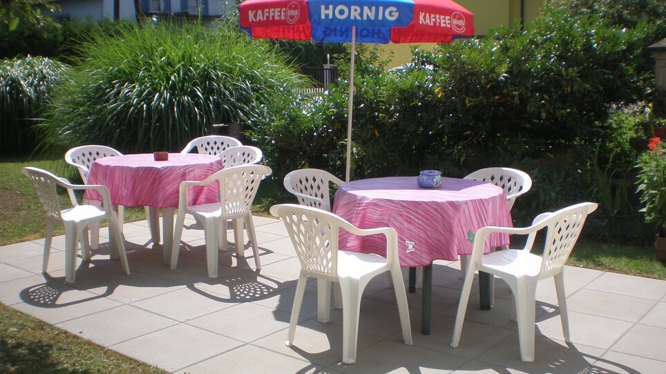 Area giardino accogliente con tavoli e sedie sotto un ombrello. | © Appartement Anne-Marie