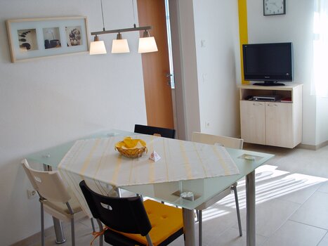 Bright dining room featuring a glass table and TV in background. | © App Alpha
