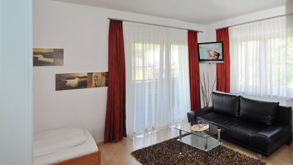 Soggiorno luminoso con divano, tavolo e TV, circondato da tende. | © Steinbichler