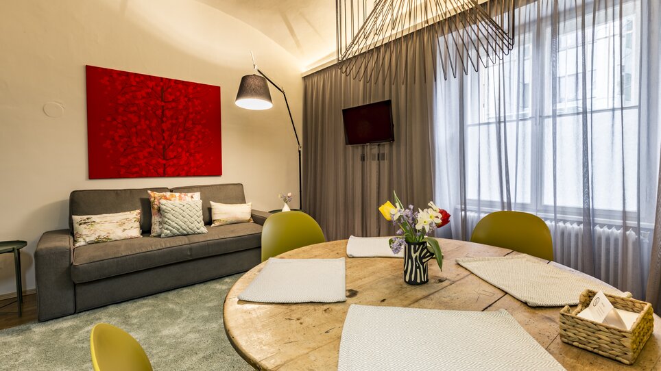 Un soggiorno con divano, tavolo e fiori. Di fronte a una parete finestrata con tende. | © GrazRentals