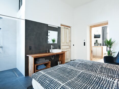 A room featuring a shower, sink, and a bed in the foreground. | © Grocholl