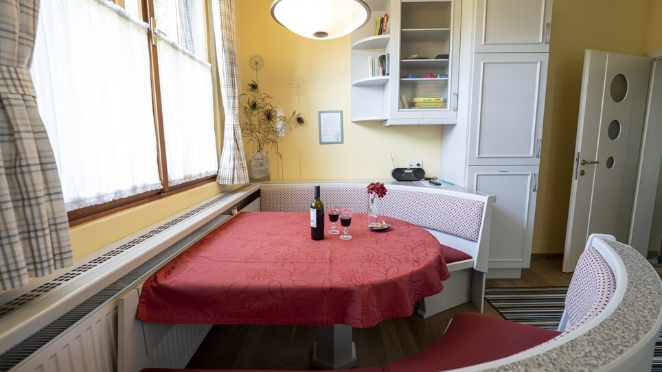 Cucina con tavolo rosso, bottiglia di vino e bicchieri. | © Haus Anna – Apartments im Garten