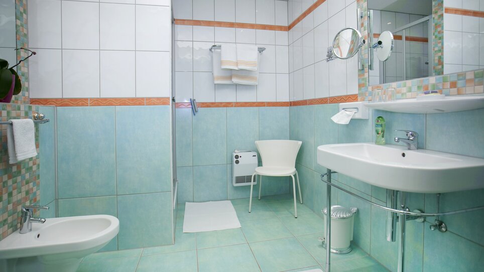 Vista luminosa del bagno con lavabo, sedia e doccia. | © Haus Anna – Apartments im Garten