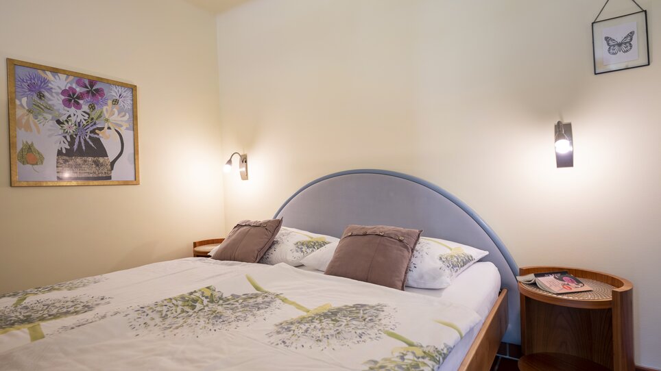 Una camera da letto con letto matrimoniale, illuminazione moderna e motivi floreali sulla biancheria. | © Haus Anna – Apartments im Garten