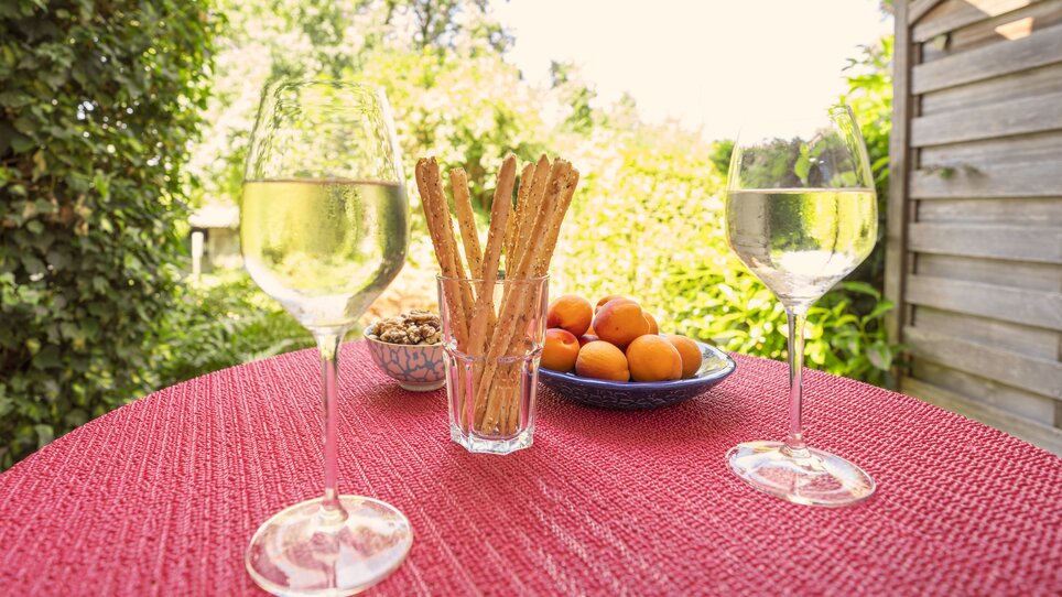 Due bicchieri di vino bianco, grissini, noci e albicocche su un tavolo in giardino. | © Haus Anna – Apartments im Garten