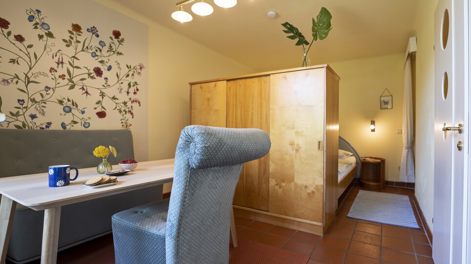 Uno spazio abitativo con tavolo, sedia, armadio e parete fiorita. | © Haus Anna – Apartments im Garten