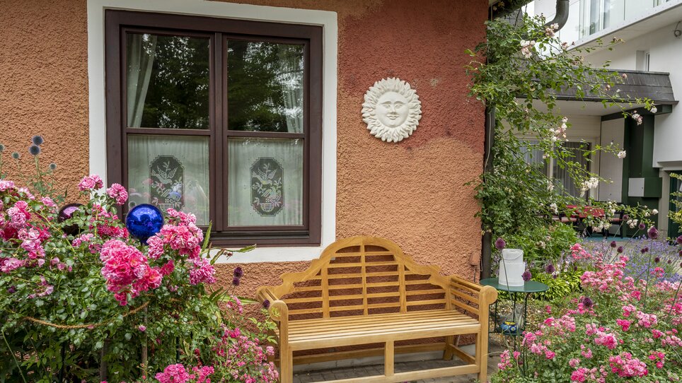 Una panchina di legno davanti a una finestra con fiori e una decorazione a simbolo del sole. | © Haus Anna – Apartments im Garten