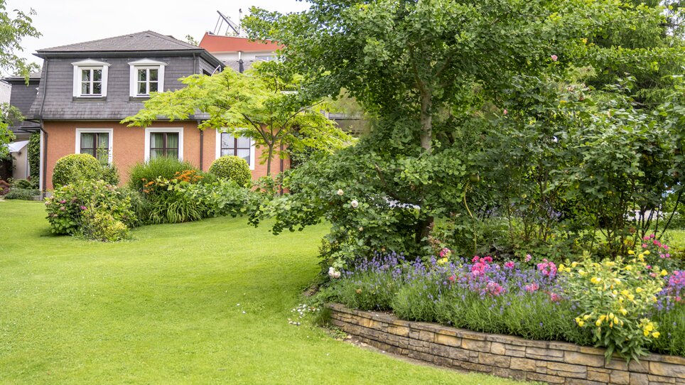 Giardino con aiuola di fiori e Haus Anna sullo sfondo. | © Haus Anna – Apartments im Garten