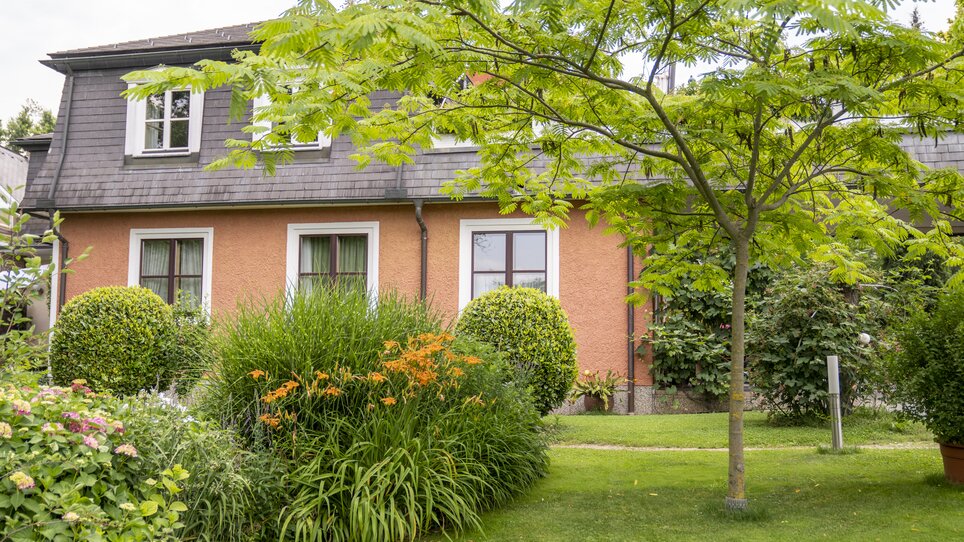 Una casa con paesaggio di giardino e alberi. Presenta finestre con tende verdi. | © Haus Anna – Apartments im Garten