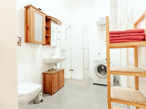 Ein helles Badezimmer mit Dusche, Waschmaschine und Holzregal. | © Lilians Apartments
