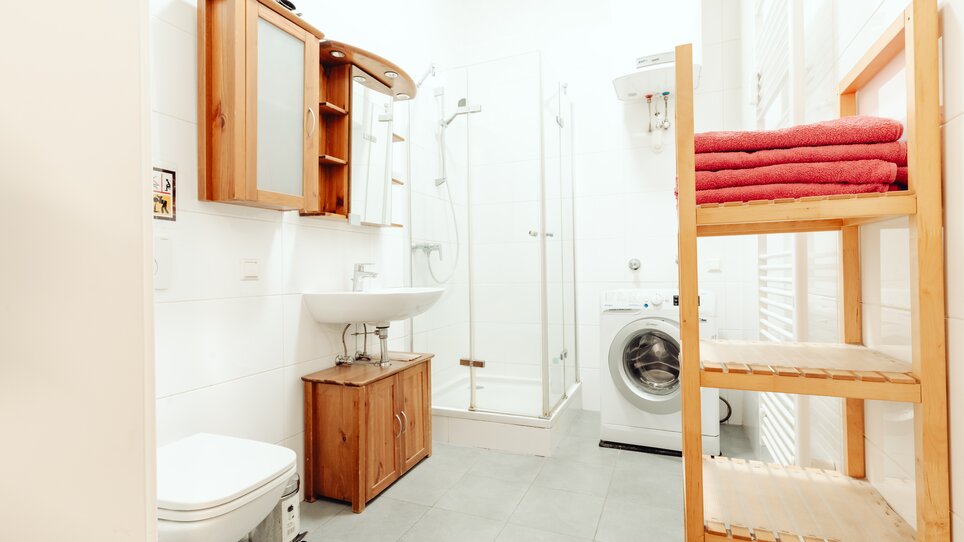 Ein helles Badezimmer mit Dusche, Waschmaschine und Holzregal. | © Lilians Apartments