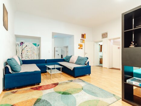 Ein modernes Wohnzimmer mit blauem Sofa, Glastisch und buntem Teppich. | © Lilians Apartments