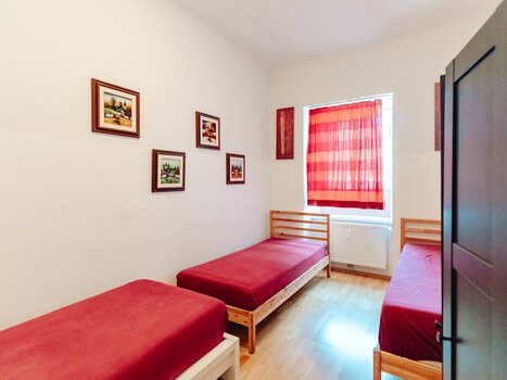 Ein helles Zimmer mit drei Einzelbetten, roter Bettwäsche und Bildern an der Wand. | © Lilians Apartments